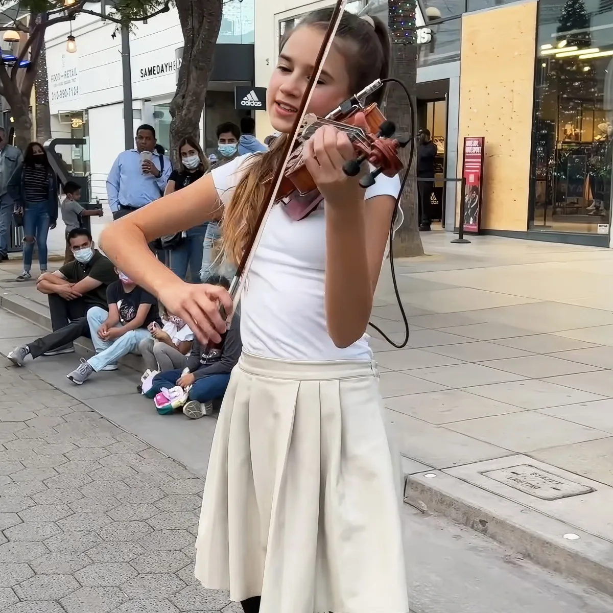 A young woman plays the violin on a city sidewalk, surrounded by onlookers and storefronts.