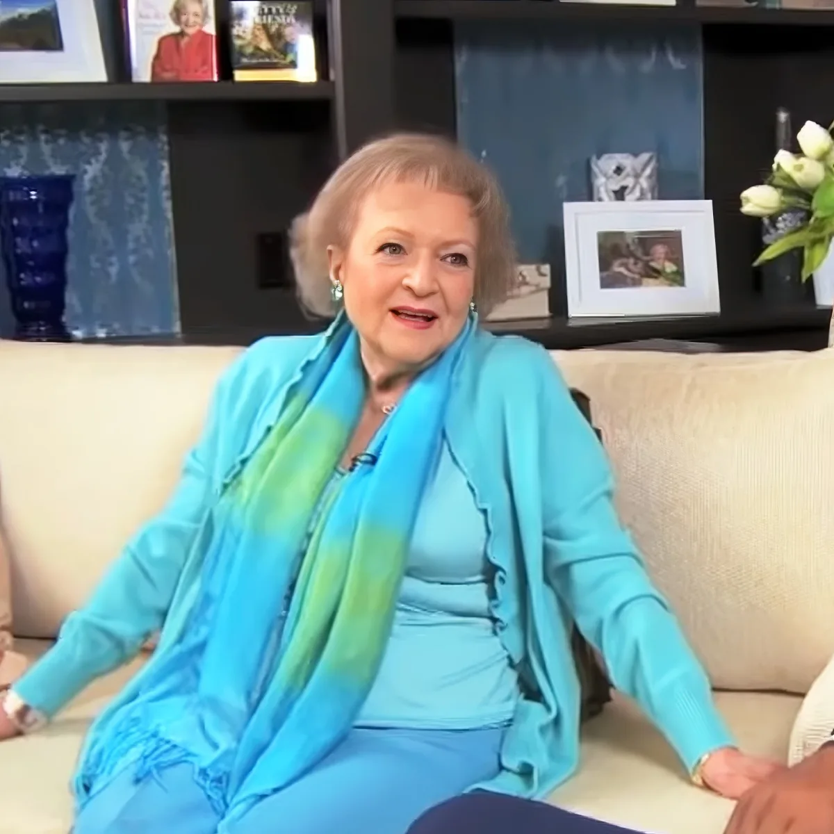 An older woman in a blue outfit and scarf sits on a beige couch, surrounded by books and decor.