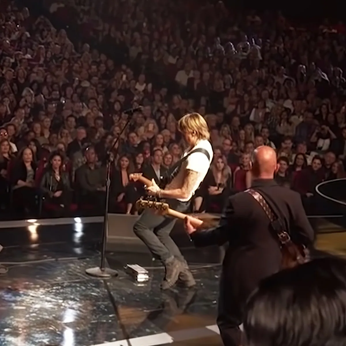 A guitarist in a white shirt and jeans plays on stage, surrounded by a large audience.
