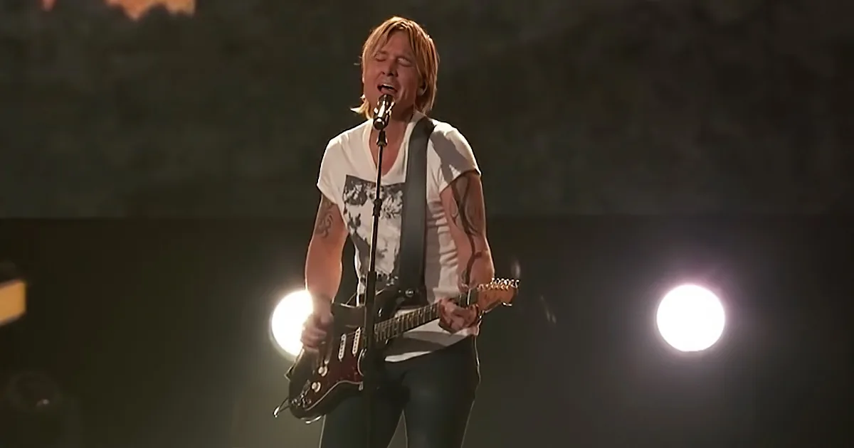 Keith Urban sings passionately on stage with a guitar, illuminated by spotlights.