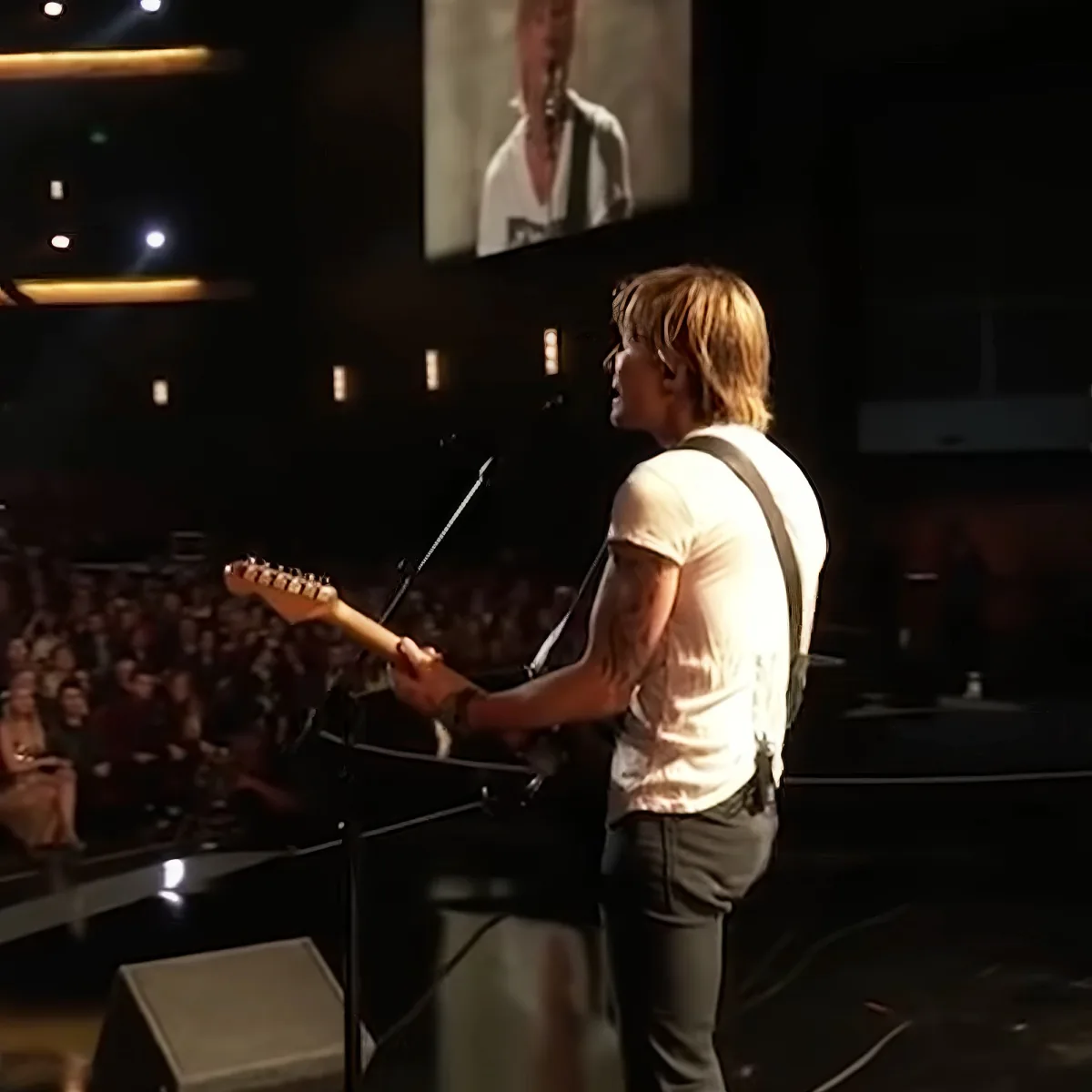 A man in a white shirt plays guitar on stage, with a large audience and a screen showing him in the background.