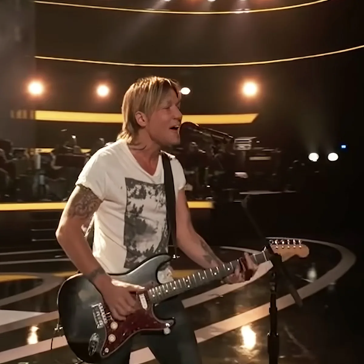 A person with tattoos plays a guitar on stage, singing into a microphone.
