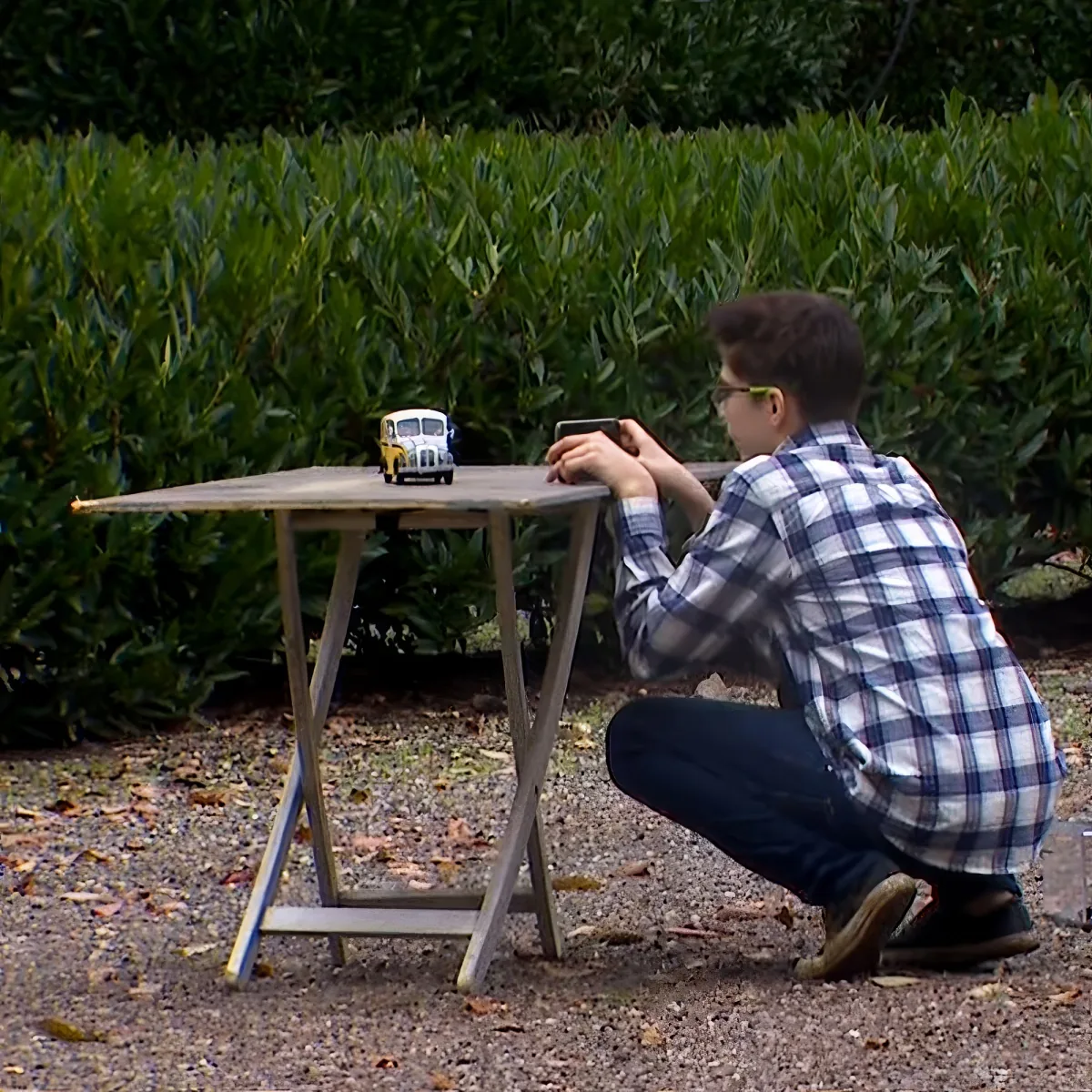 A boy in a plaid shirt crouches to take a photo of a toy bus on a small table.