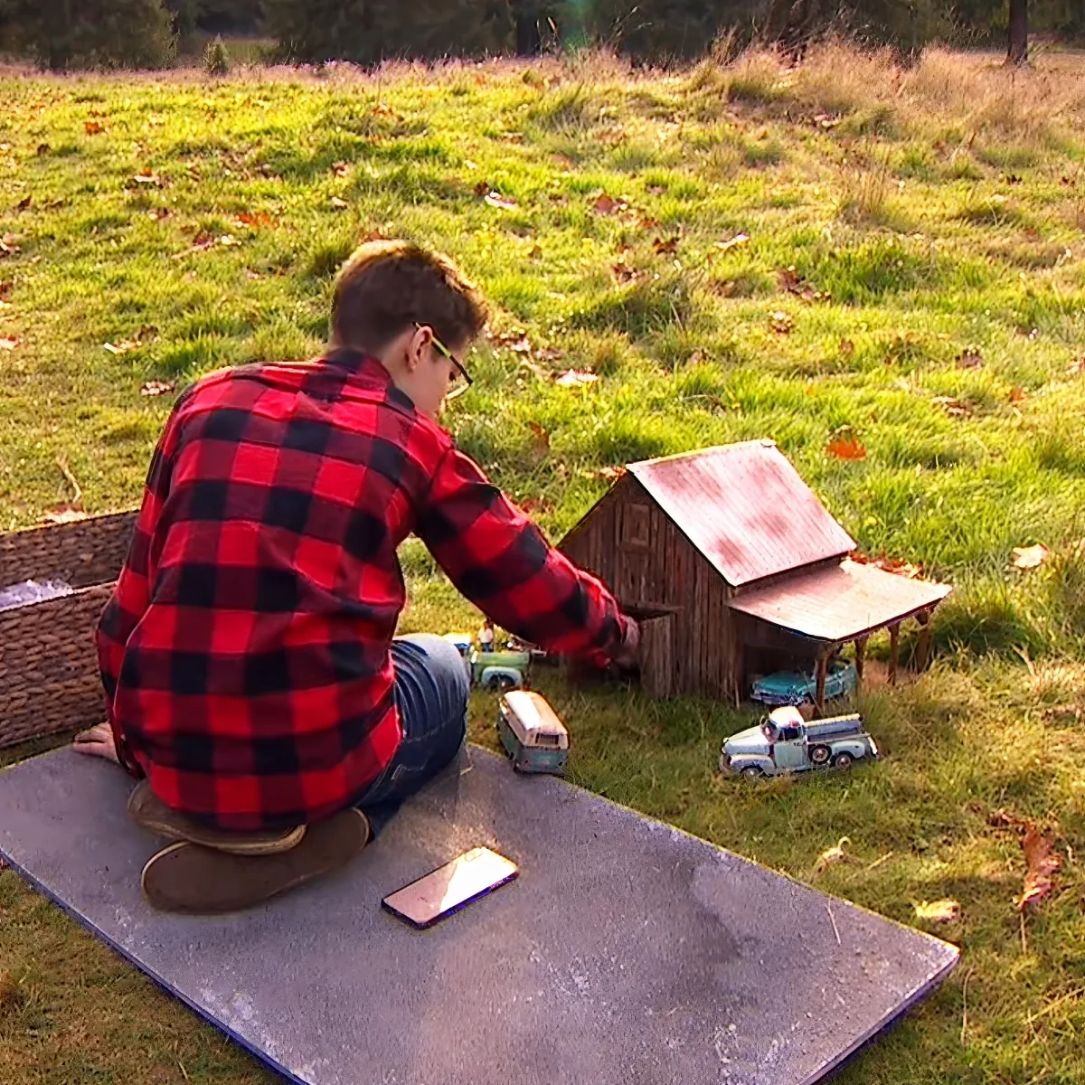 A boy in a red plaid shirt sits on a concrete slab, interacting with a miniature barn and toy vehicles in a grassy field.