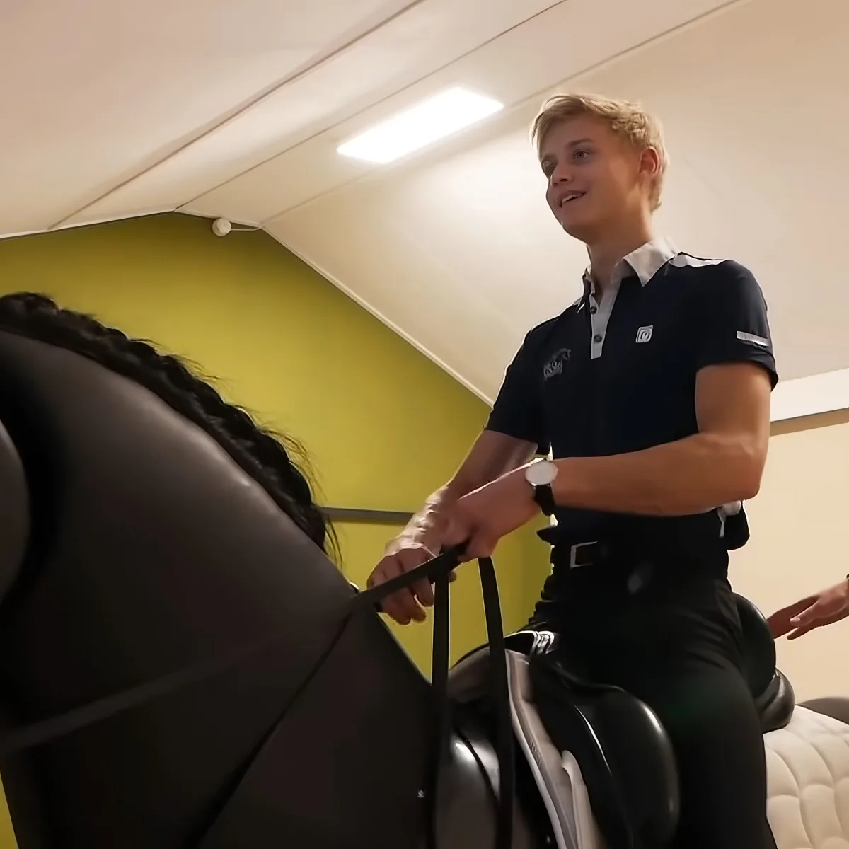 A man in a dark polo shirt and black pants is riding a stationary exercise bike in a room with a green wall.