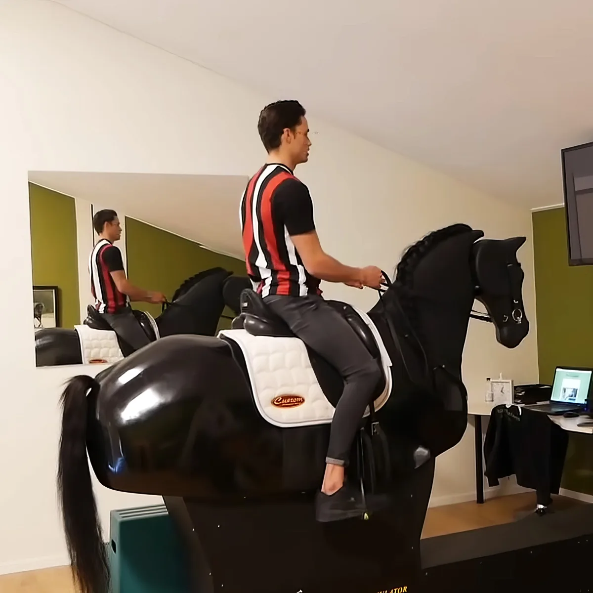 A man in a red and black striped shirt rides a black horse simulator in a room with a green wall and a mirror.