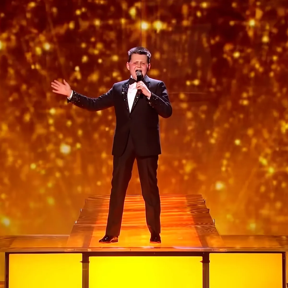 A man in a black suit sings on stage with a microphone, arms outstretched, against a golden backdrop.