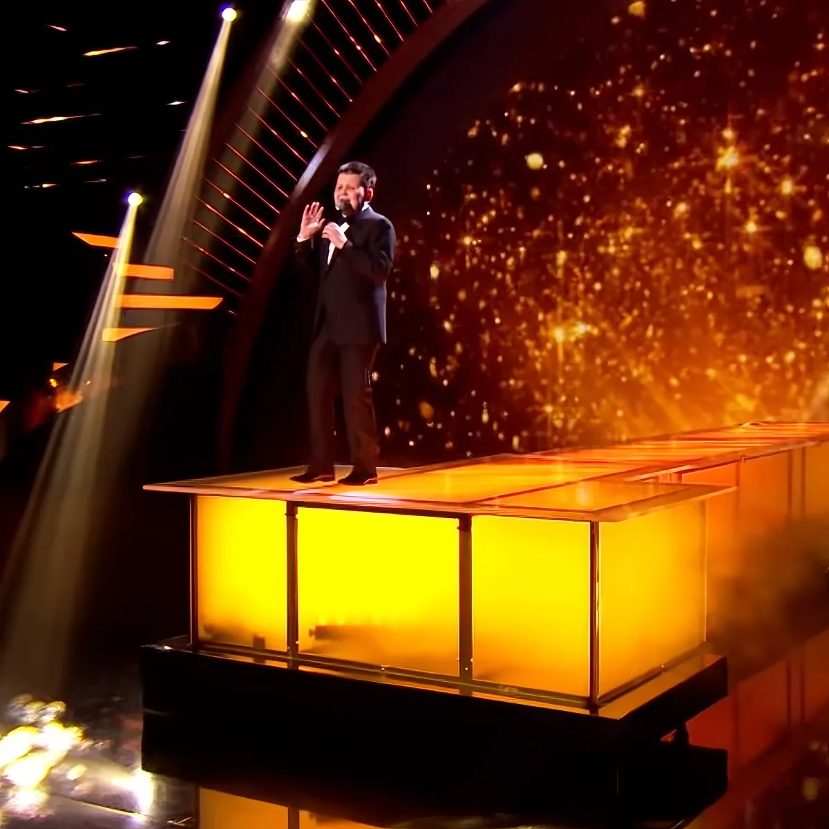 A man in a suit stands on a glowing platform, illuminated by stage lights and a golden backdrop.
