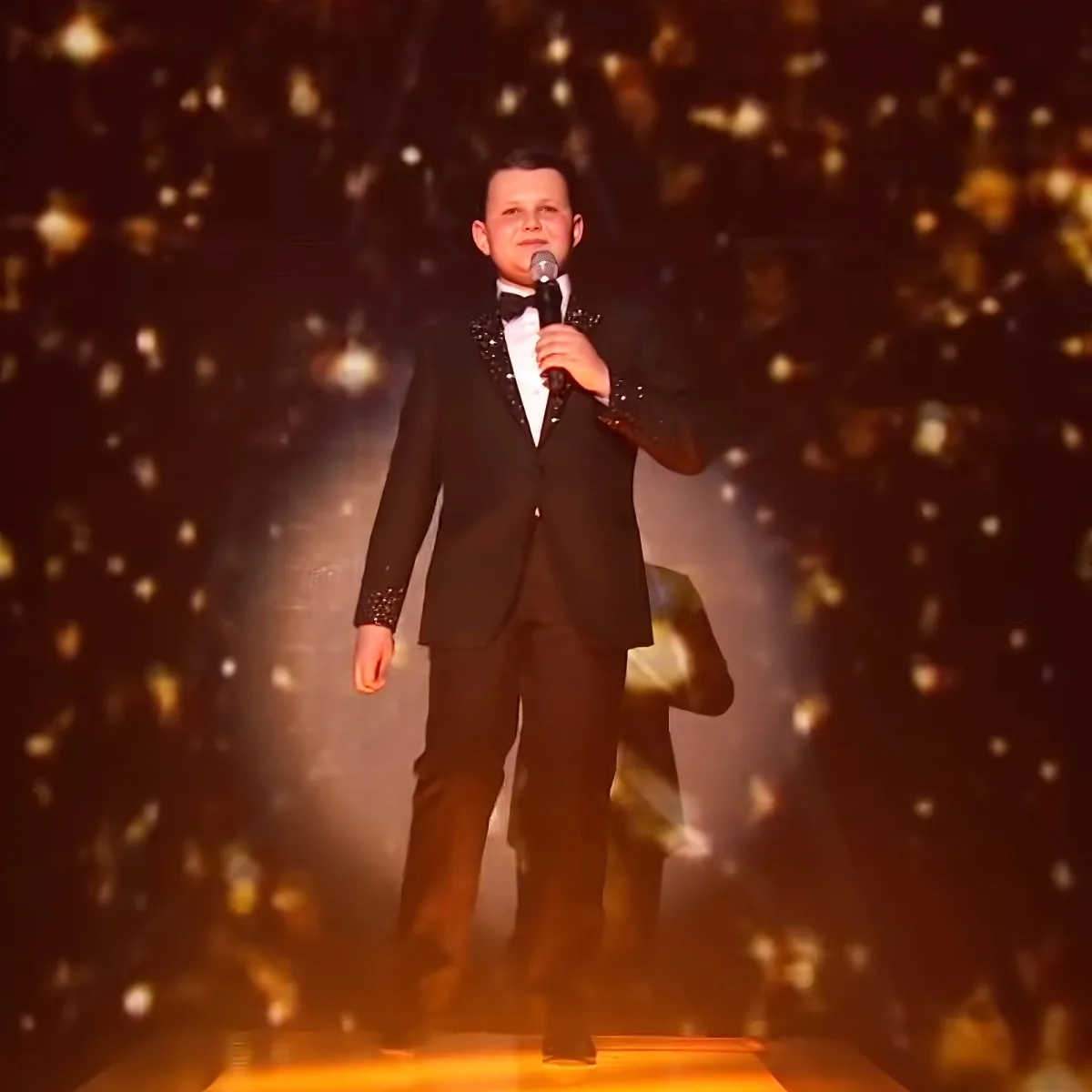 A young boy in a black suit holds a microphone on stage amidst golden confetti.