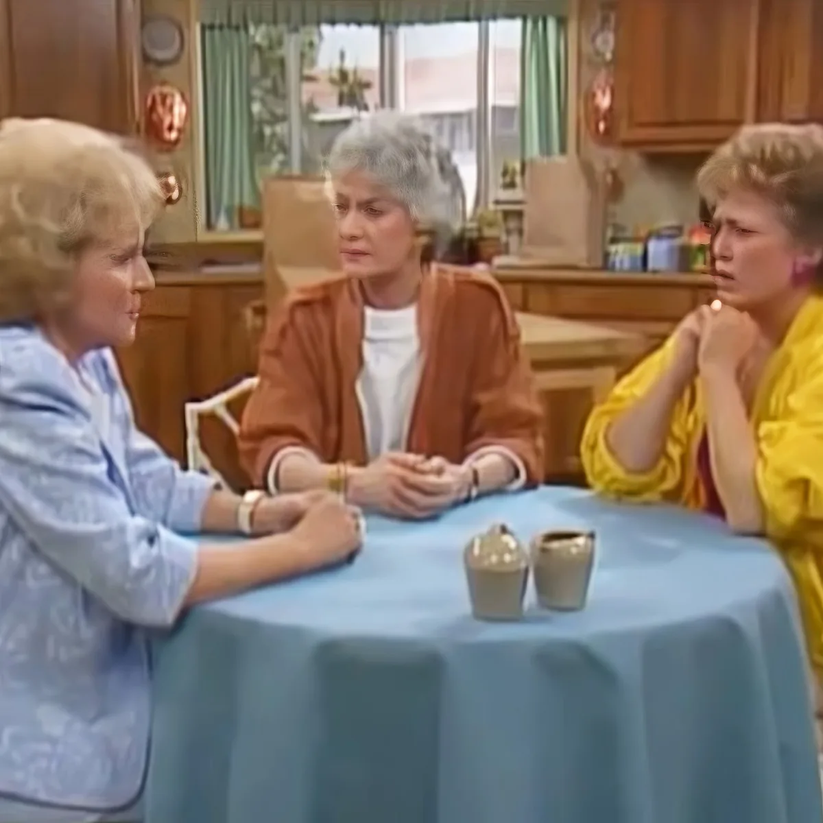 Three women sit at a blue table in a kitchen, engaged in conversation.