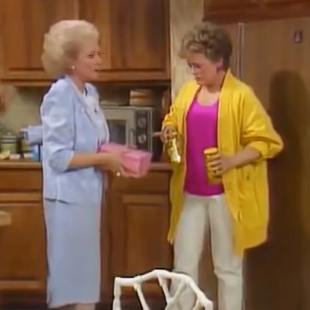 Two women in a kitchen, one in a blue suit holding a pink object, the other in a yellow jacket holding a drink.