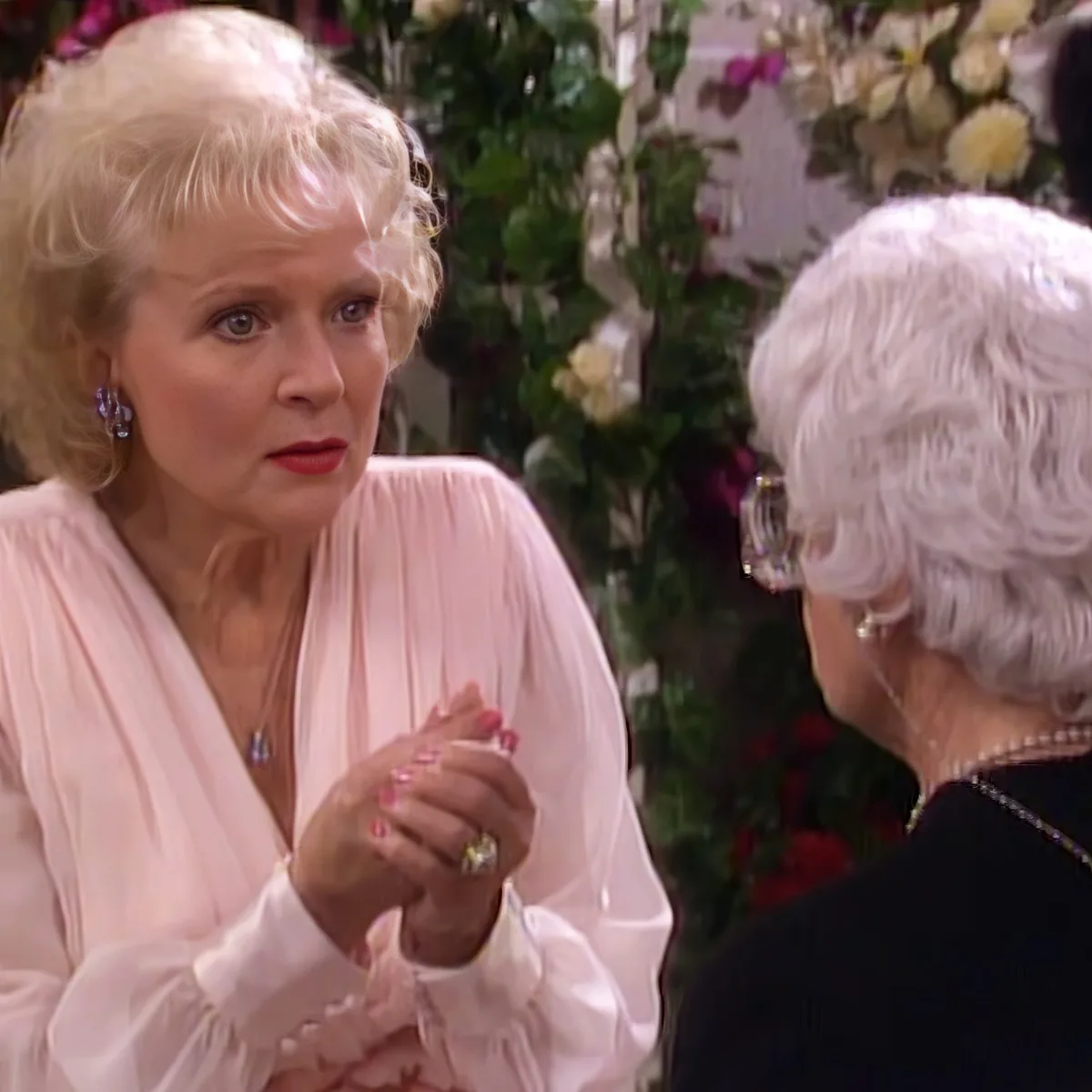 An older woman in a pink blouse gestures with her hands while talking to another woman with white hair.