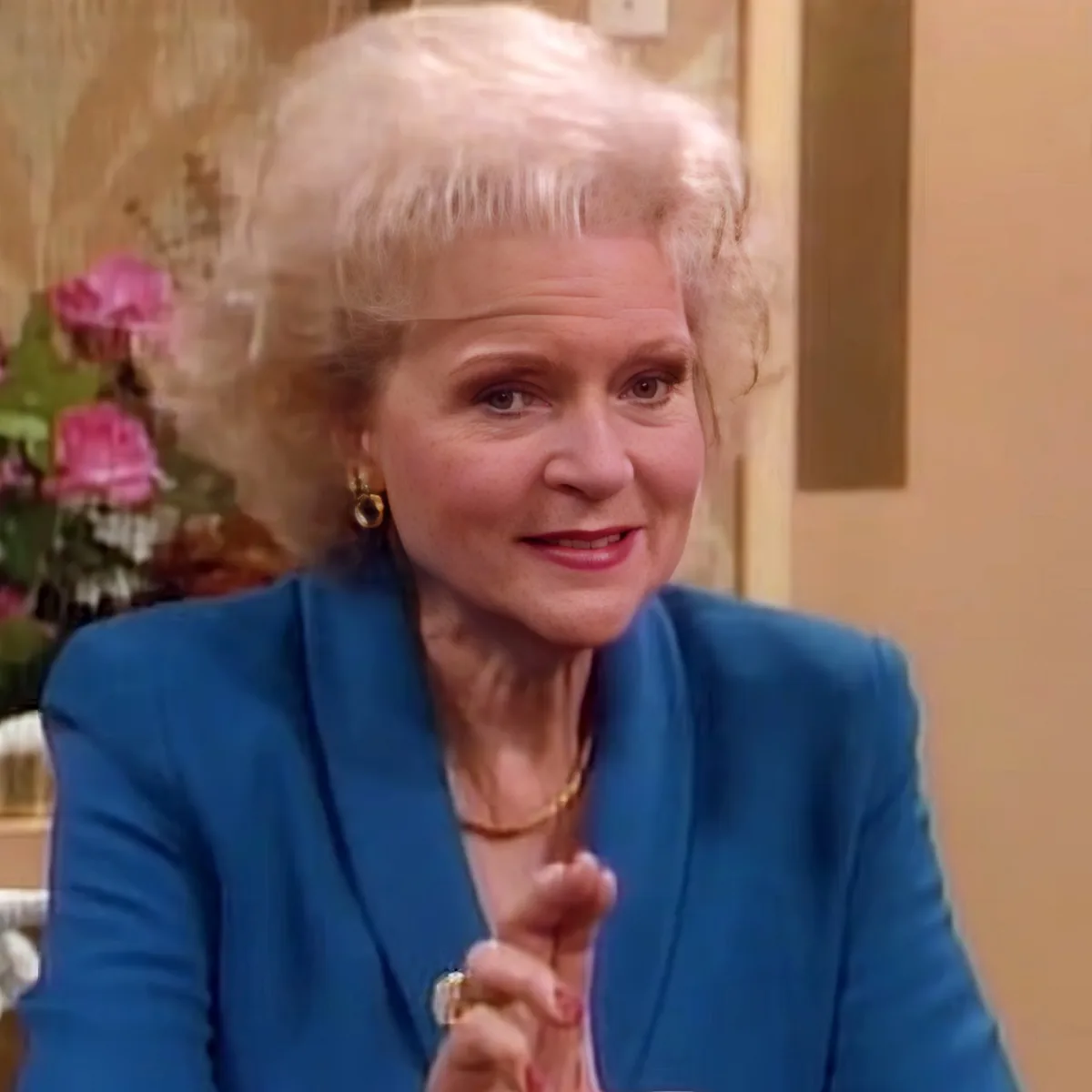 A woman with short, light-colored hair, wearing a blue blazer and a gold necklace, is gesturing with her hands.