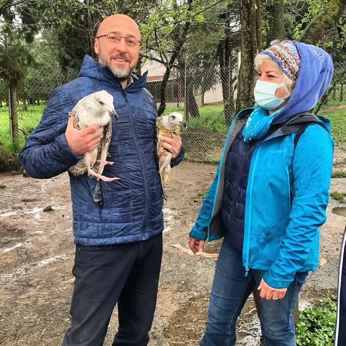 A man holds two birds, smiling, while a woman in a mask looks on in a park setting.