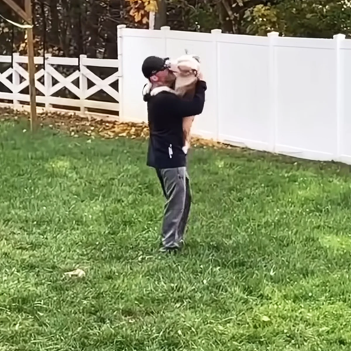 A man holds a child in a grassy yard near a white fence.
