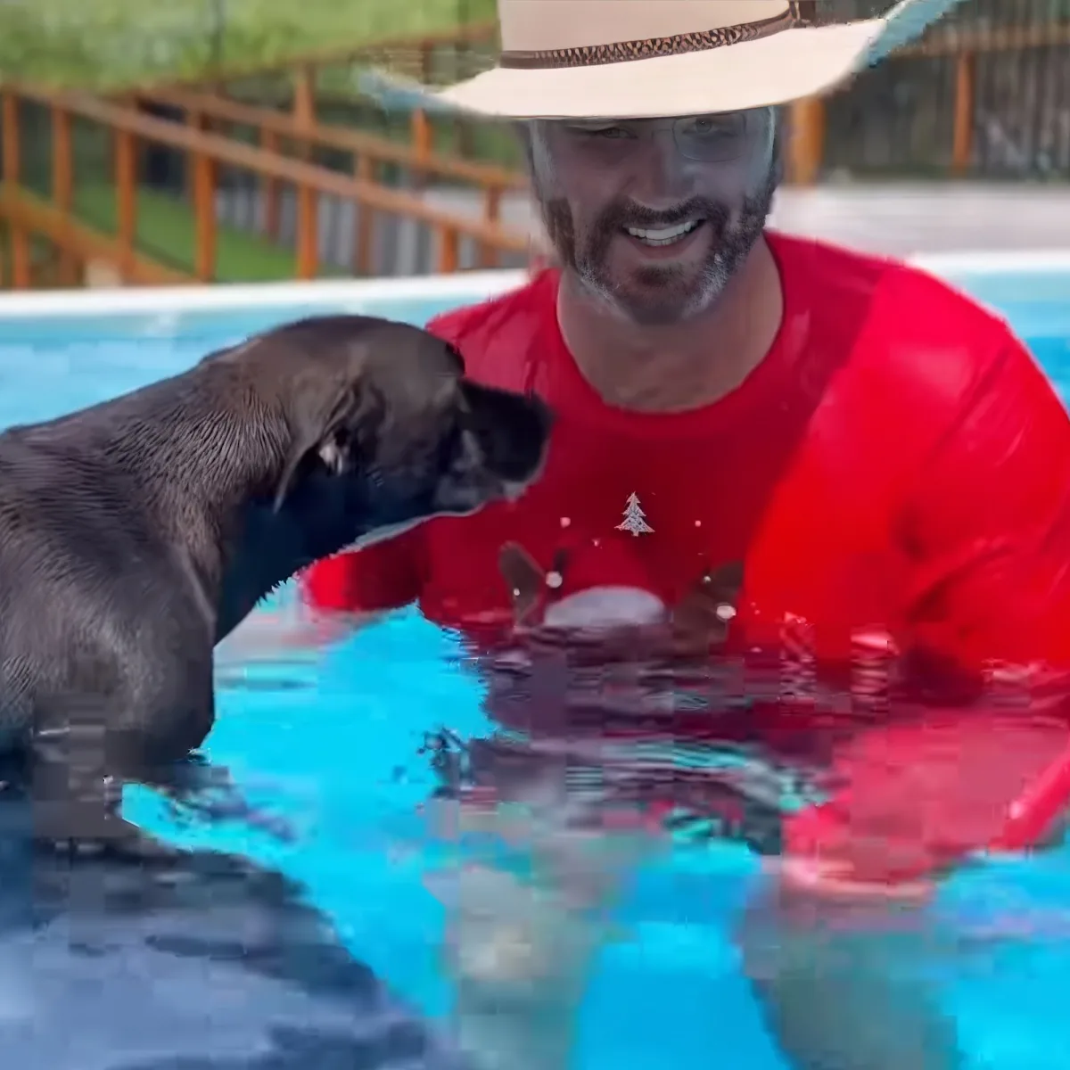 A man in a cowboy hat and red shirt is in a pool with a black puppy, both smiling and enjoying the water.