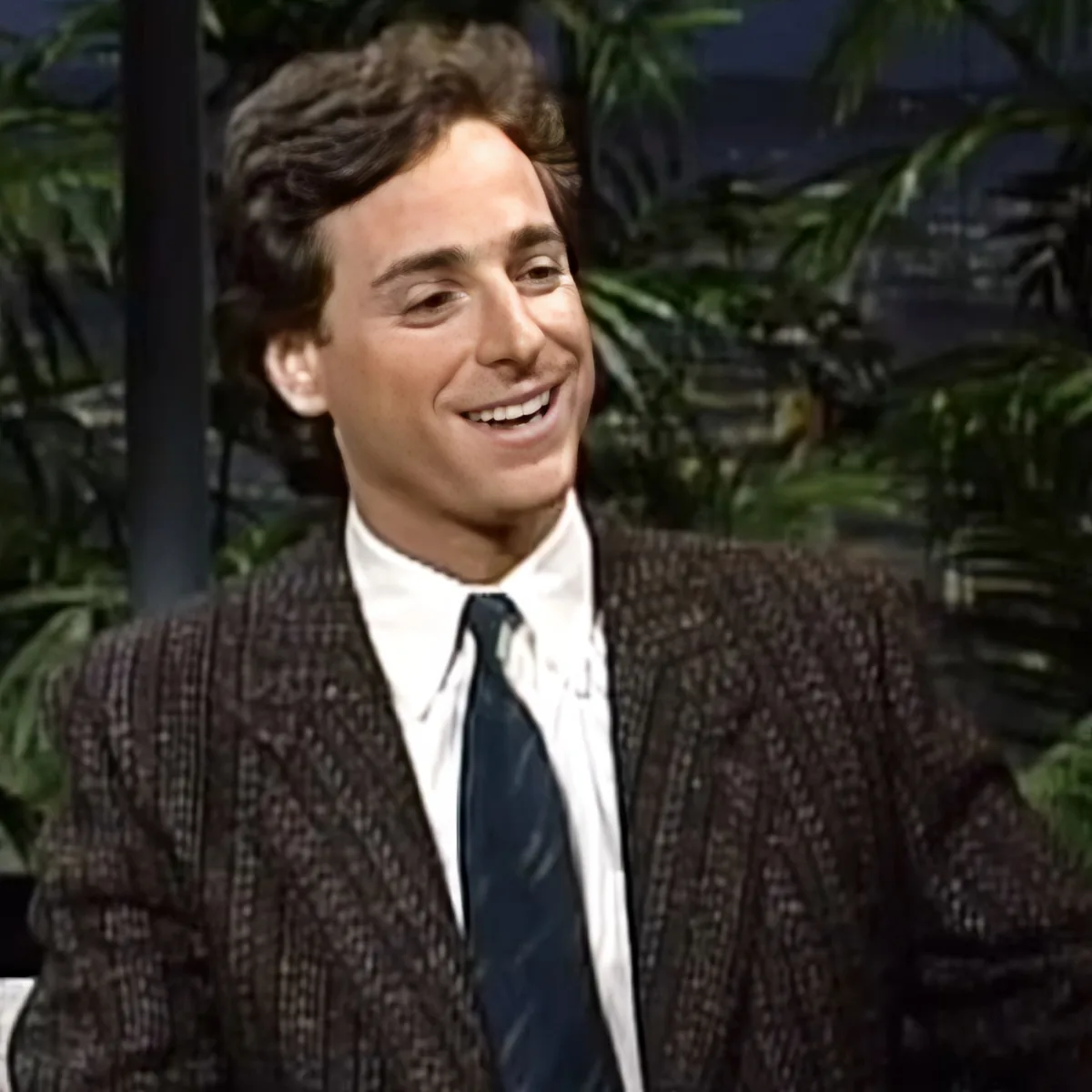 A man in a patterned suit and tie smiles, with palm trees and a dark background behind him.