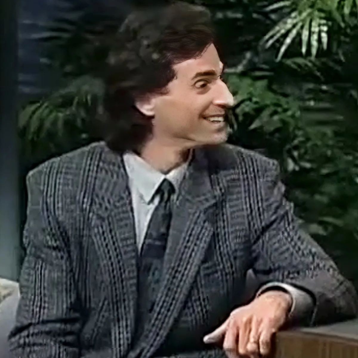 A man in a plaid suit jacket and tie is smiling, seated with a plant in the background.