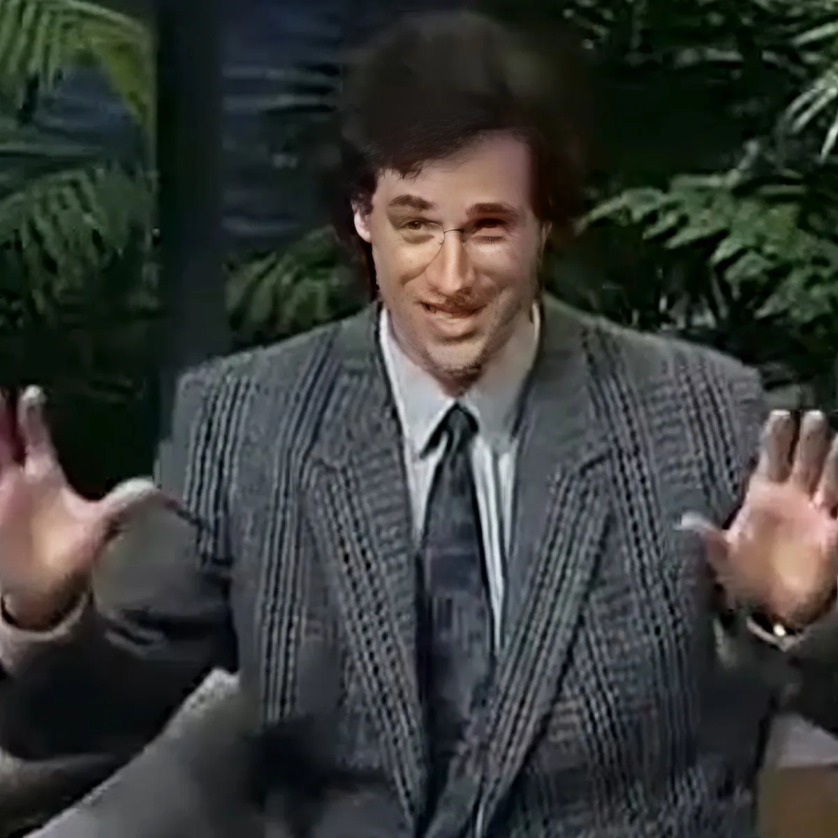 A man in a striped suit jacket and tie is smiling and gesturing with his hands.