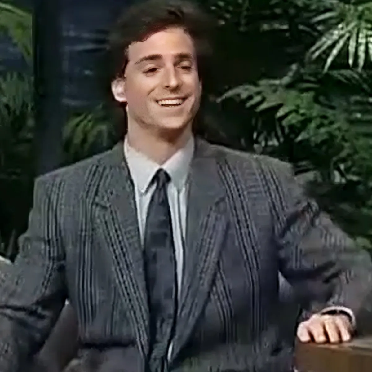A man in a striped suit and tie smiles, hands on a desk, with greenery in the background.