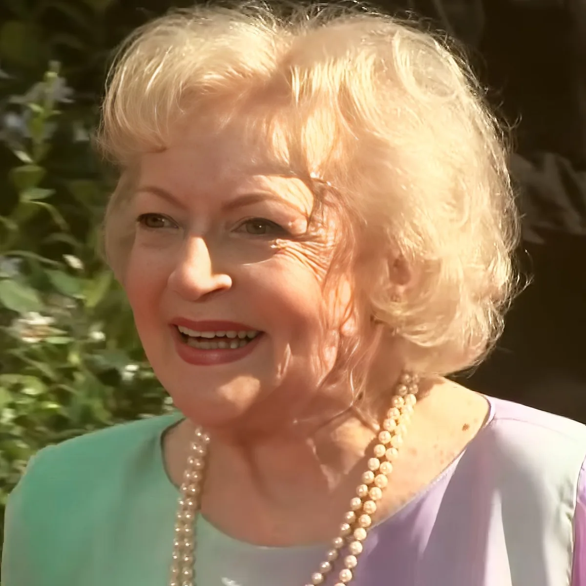 An elderly woman with short, light-colored hair, wearing a pearl necklace and a light blue top, smiles softly.