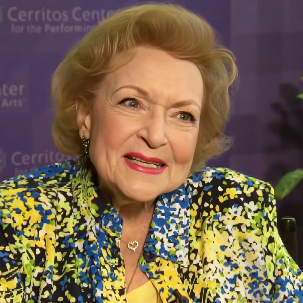 An older woman with blonde hair, wearing a colorful floral jacket, smiles while seated in front of a purple backdrop.