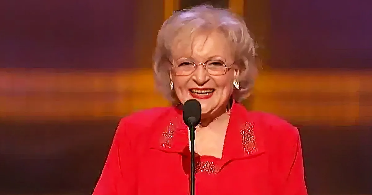 An elderly woman in a red outfit speaks into a microphone on stage.