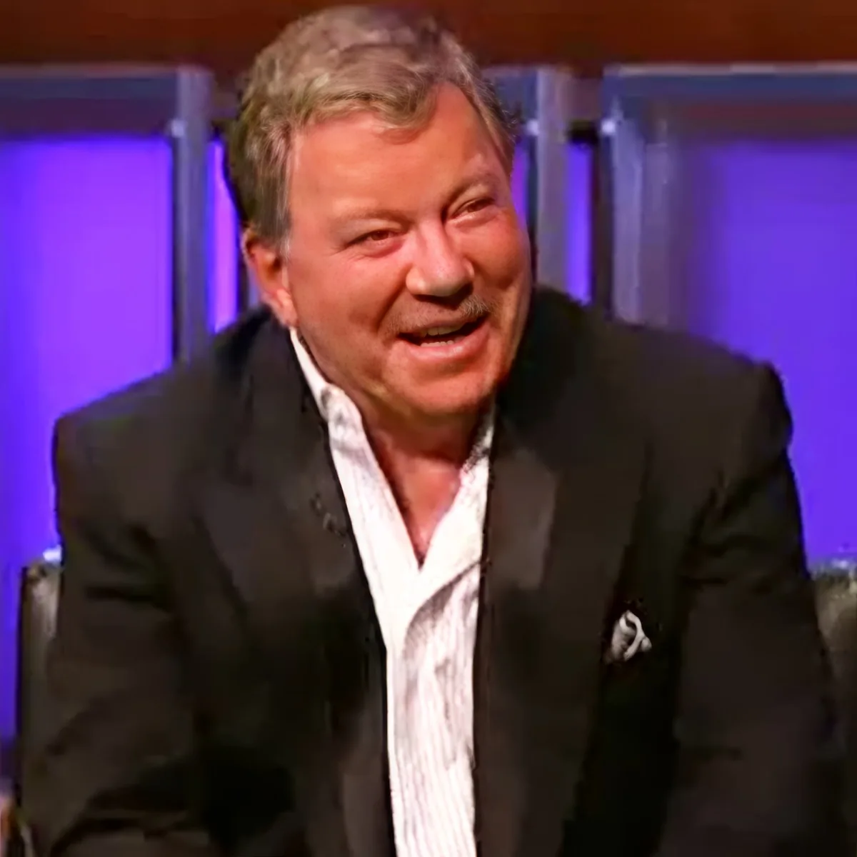 A man in a suit and striped shirt is smiling, seated in a studio setting with purple lighting.