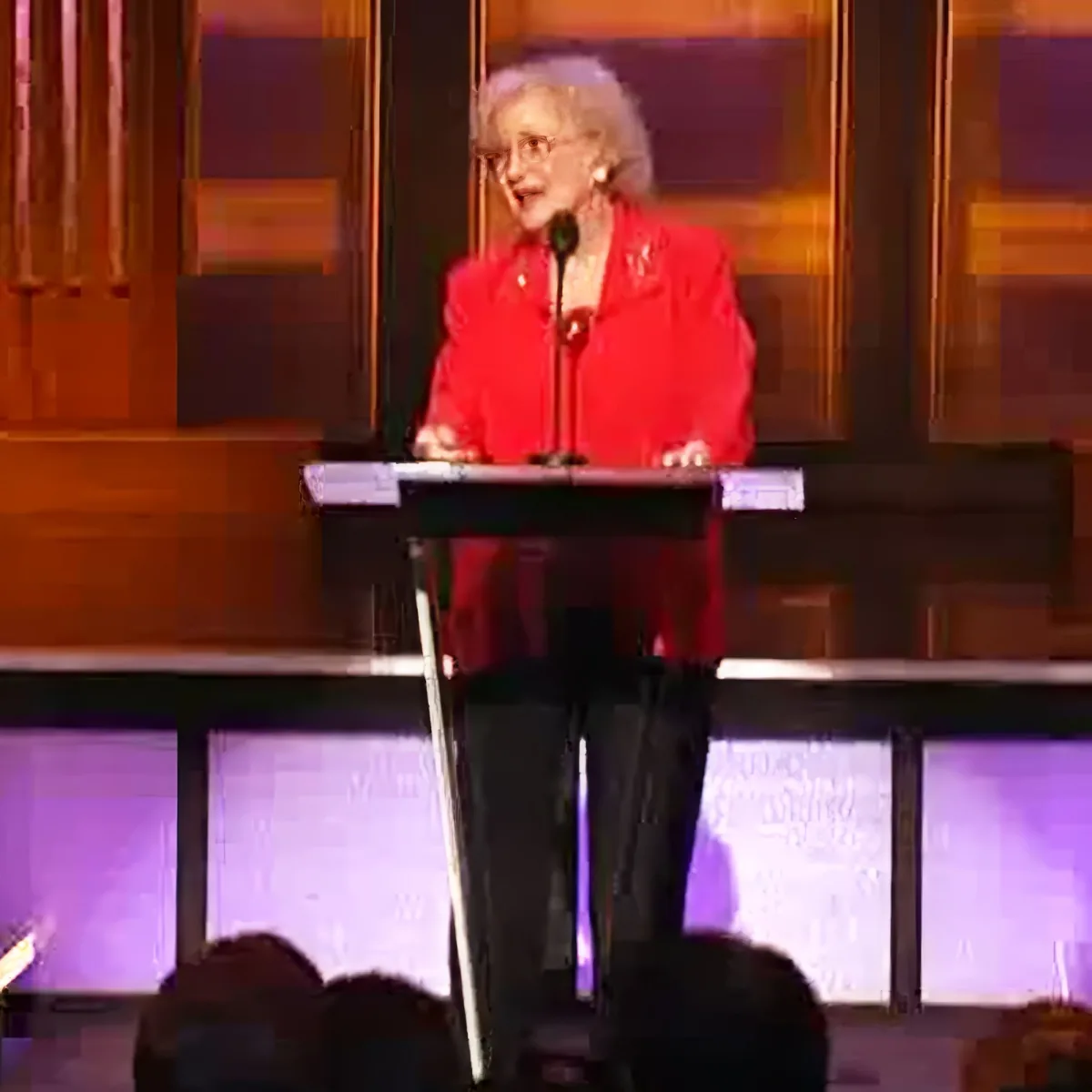 A woman in a red jacket stands at a podium, speaking into a microphone, with a wooden backdrop and purple lighting.