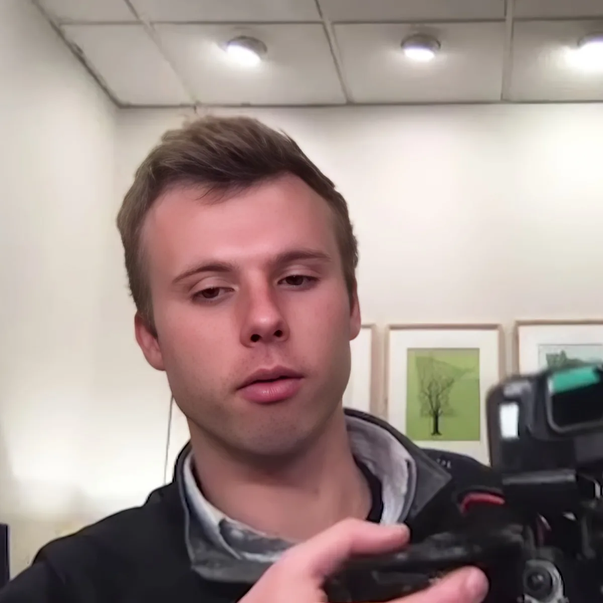 A person holding a camera, wearing a black jacket and gray shirt, in a room with framed artwork on the wall.