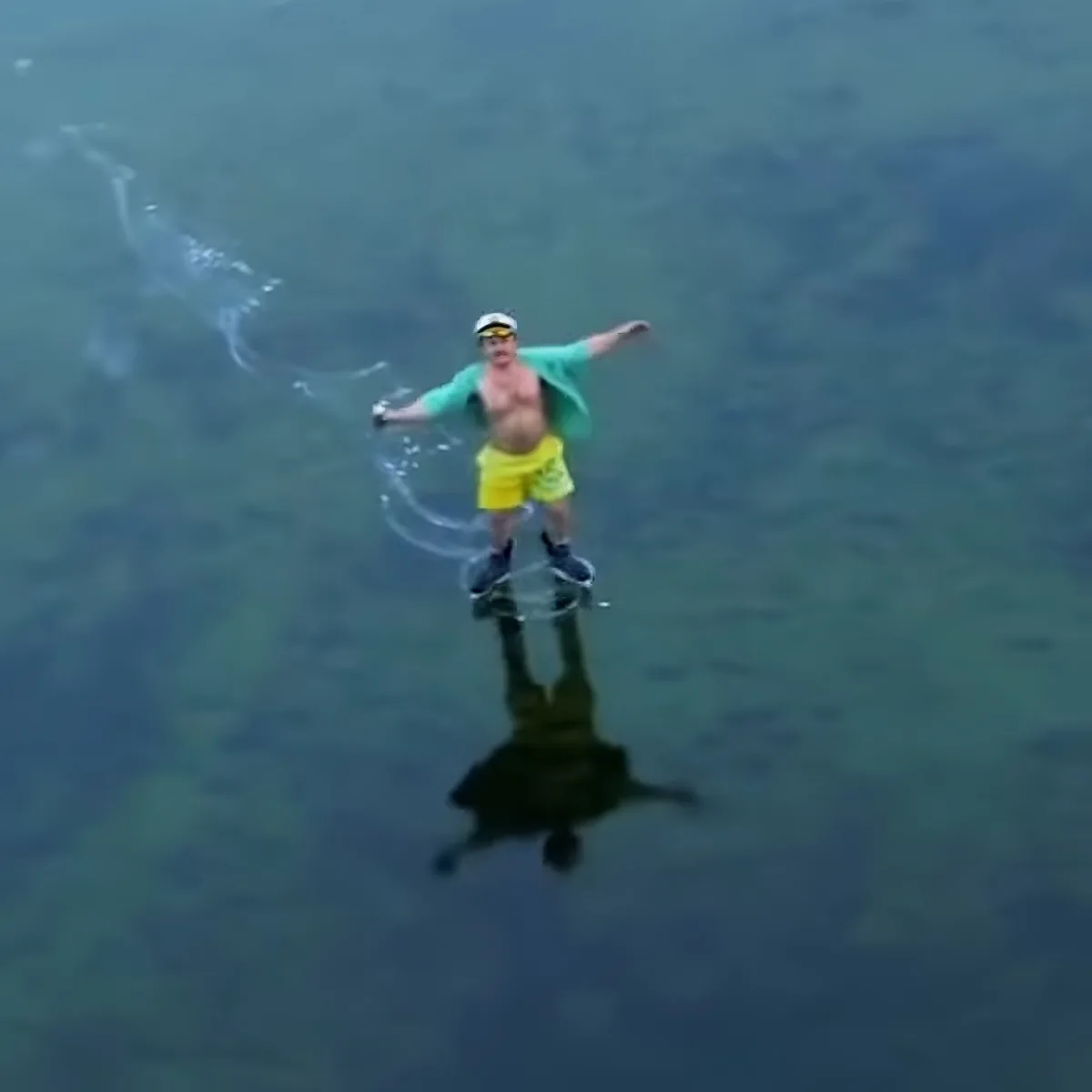 A man in yellow shorts and green shirt is standing on a glassy surface, creating ripples in the water.