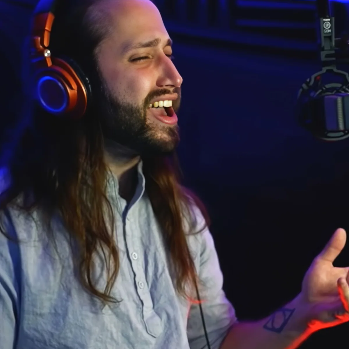 A man with long hair and beard wearing headphones, speaking into a microphone in a dimly lit room.
