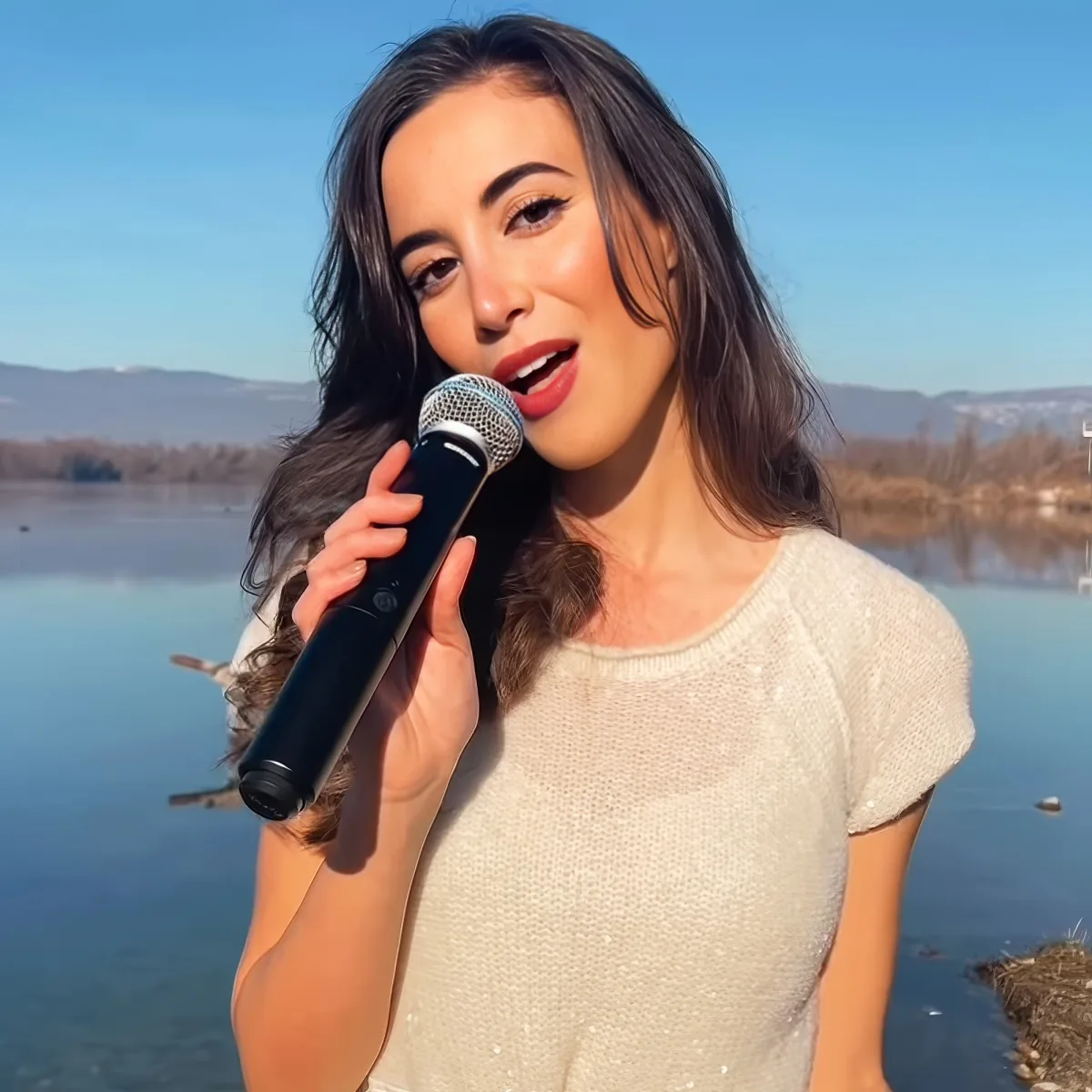 A woman holds a microphone, singing outdoors by a lake with mountains in the background.