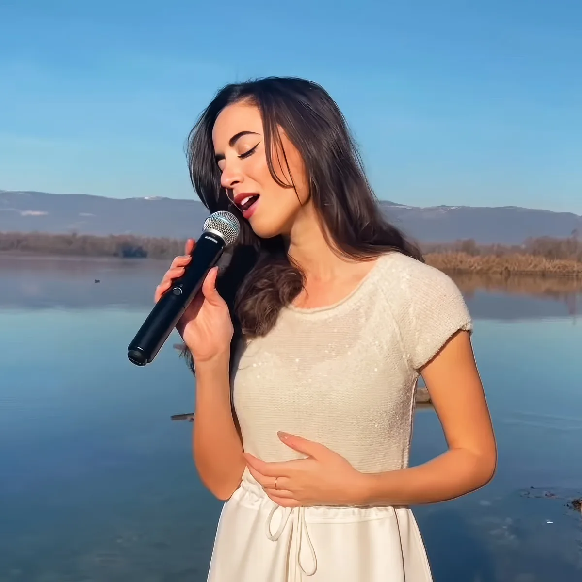 A woman in a white dress sings into a microphone by a serene lake with mountains in the background.