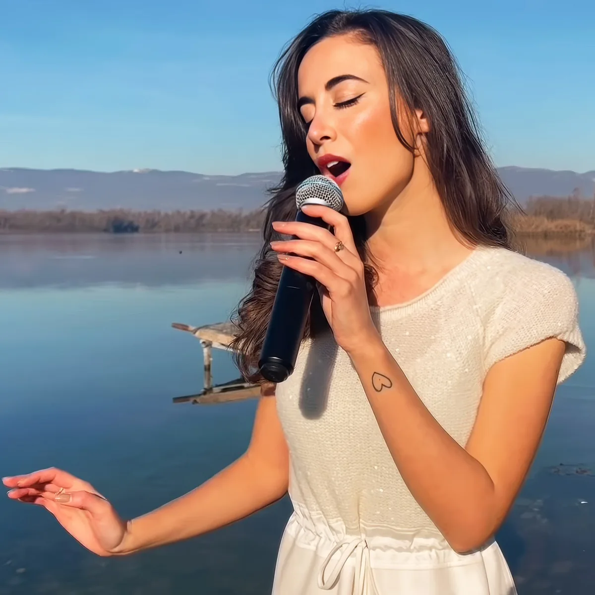 A woman in a white dress sings into a microphone by a lake with mountains in the background.