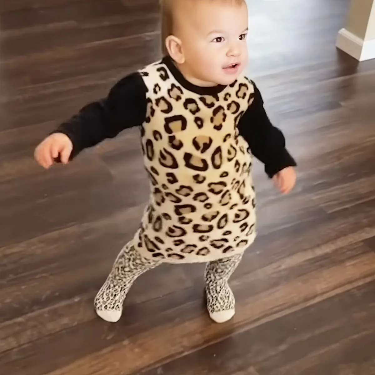 A baby in a leopard print dress and leggings is walking on a wooden floor.