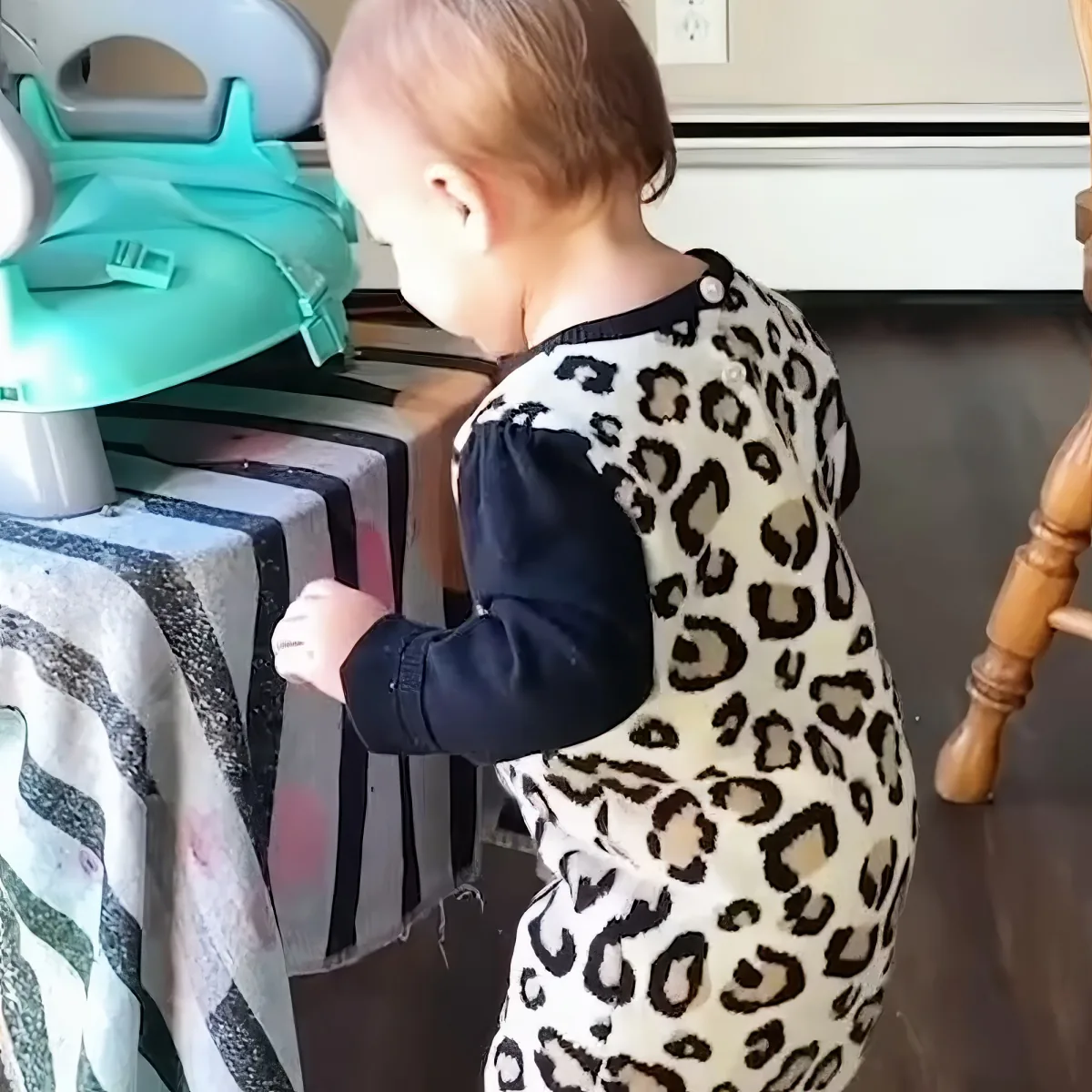 A baby in a leopard print onesie stands near a table, reaching out with one hand.