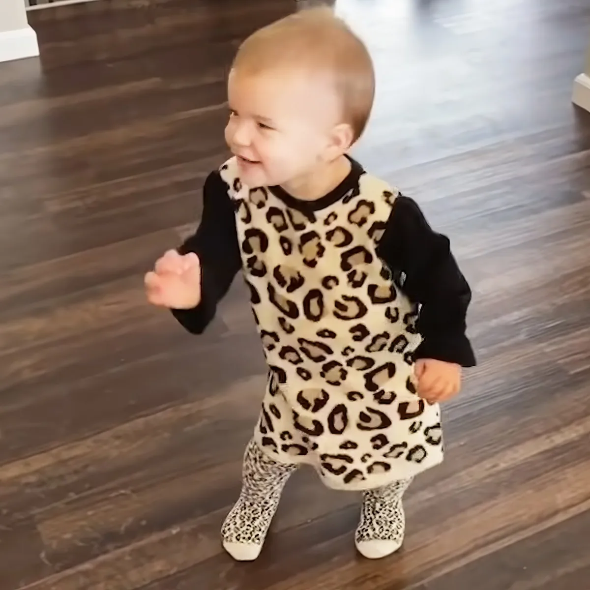 A toddler in a leopard print outfit is walking on a wooden floor, smiling.