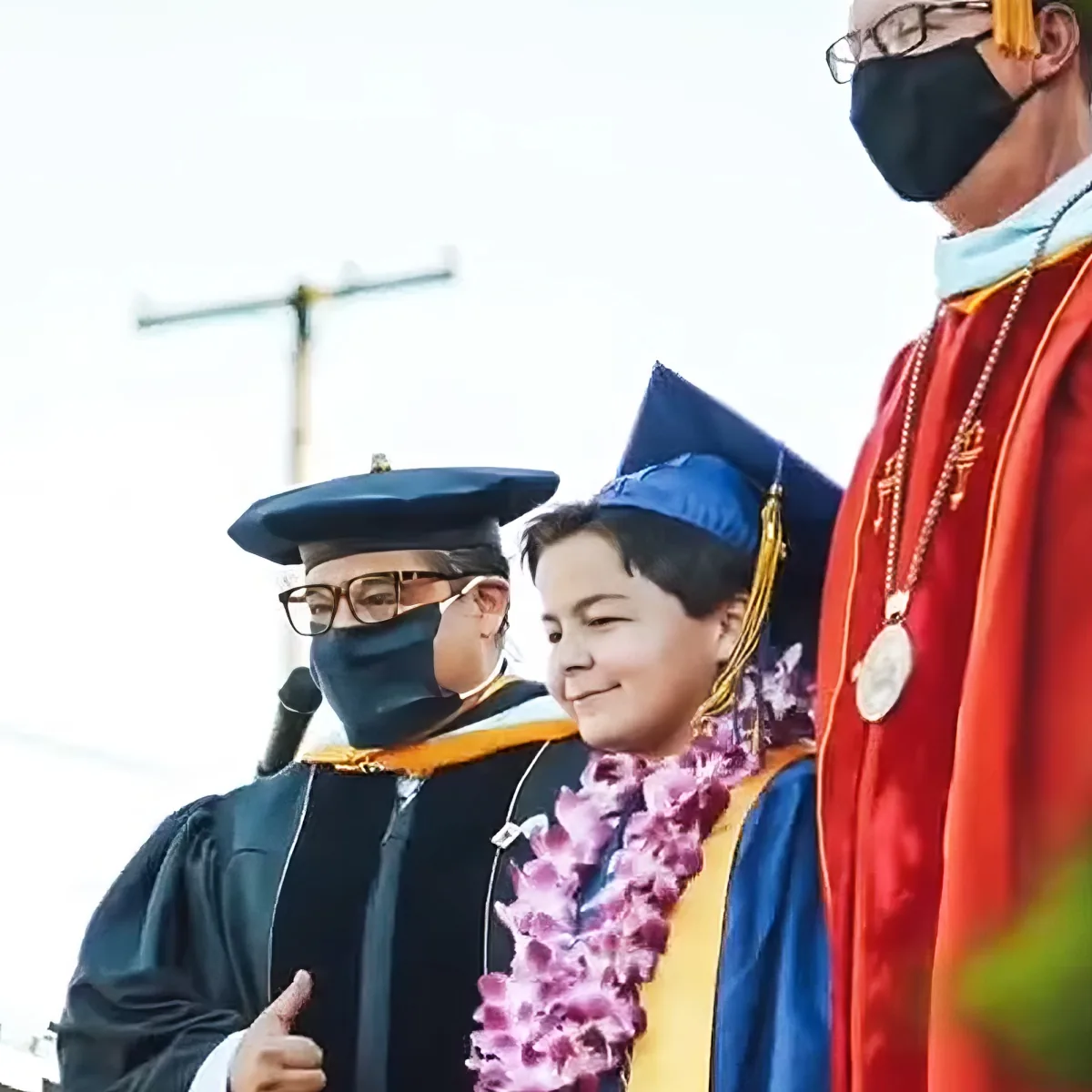 A young graduate in a blue gown and lei smiles at the camera during a graduation ceremony.