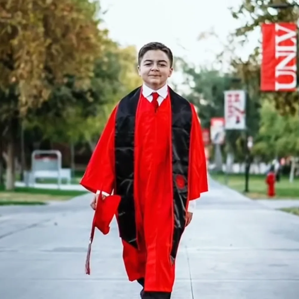 A young person in a red graduation gown and black stole walks on a campus path.