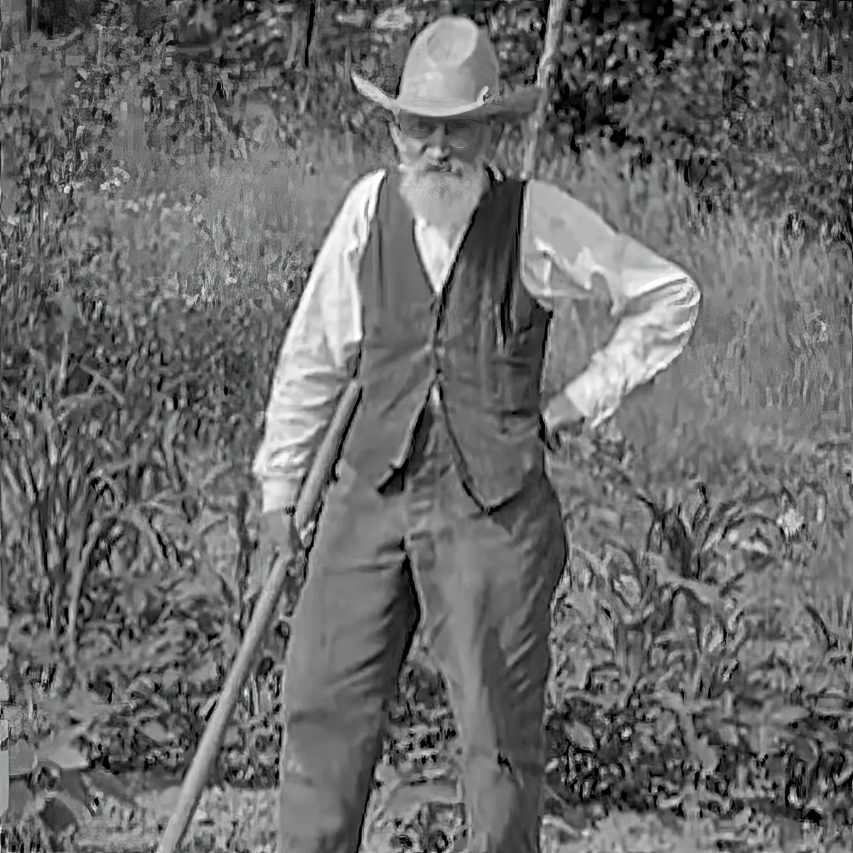 A man in a hat and vest stands in a field, holding a long object, with plants and grass around him.