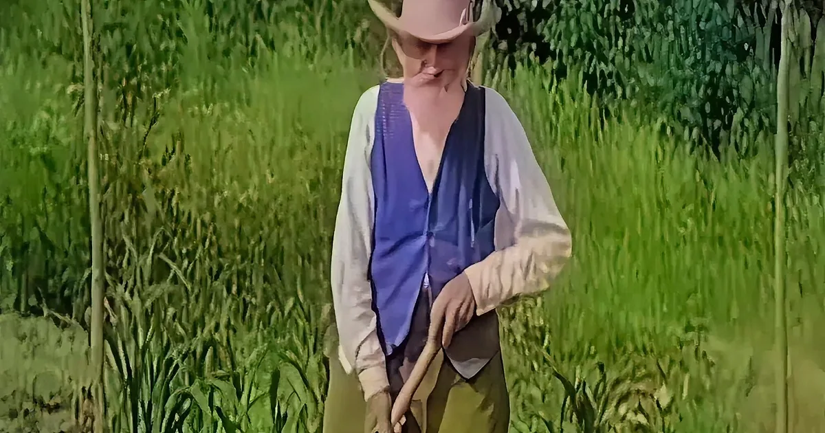 A person in a cowboy hat and vest stands in a field, holding a tool, surrounded by tall green grass.