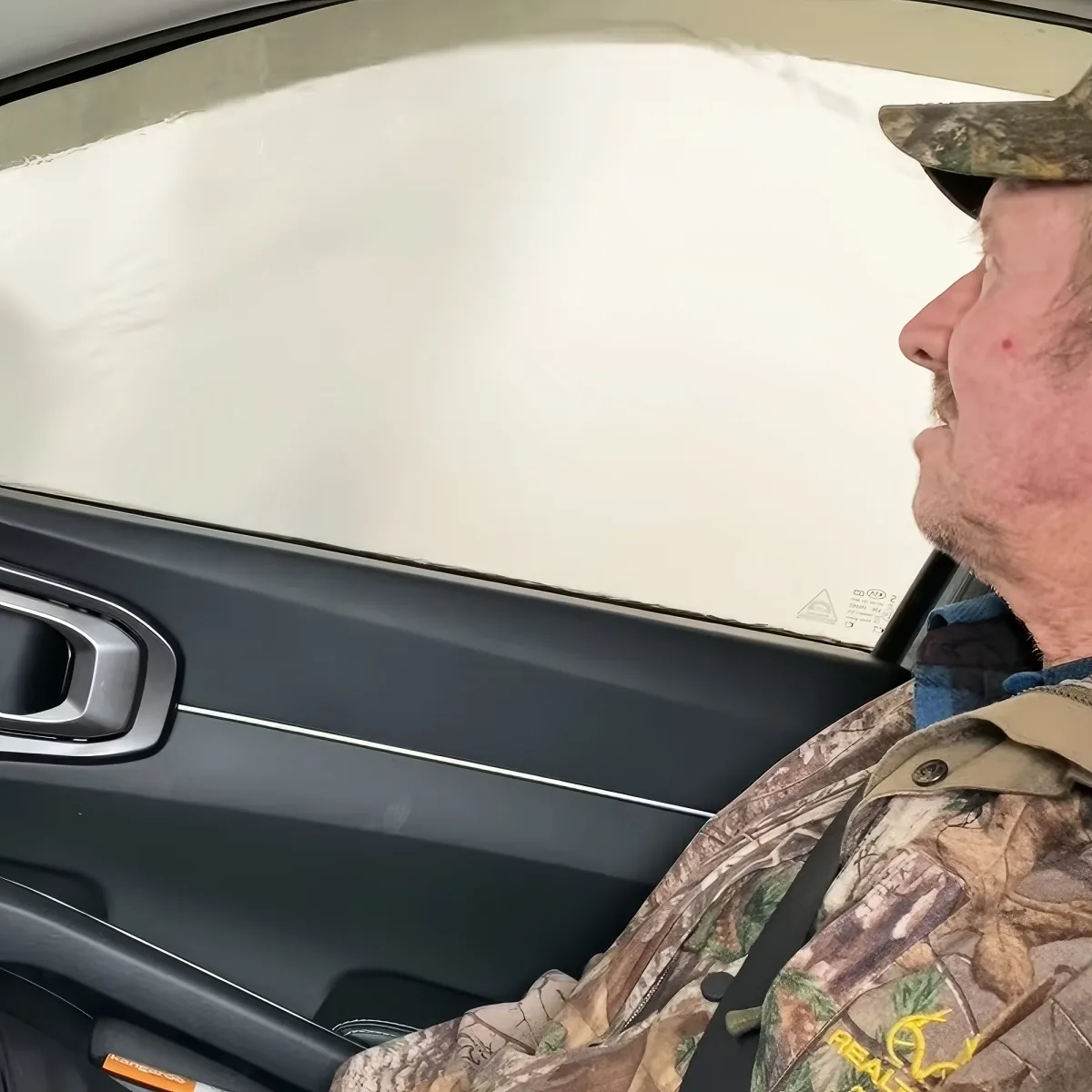 A man in camouflage attire and a hat is sitting in a car, looking out the window.
