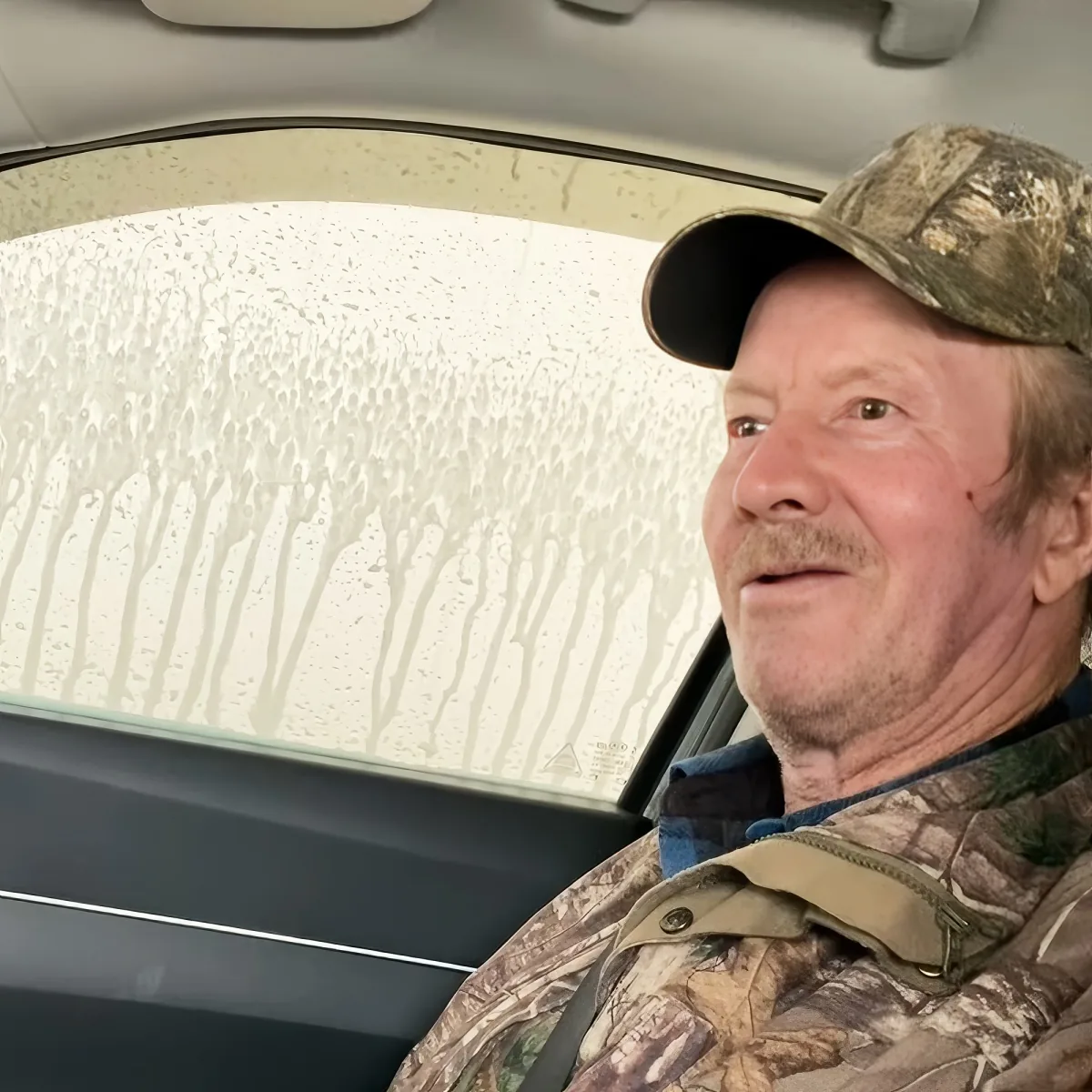 A man in camouflage attire sits in a car, looking out the window with soapy water streaks on the glass.