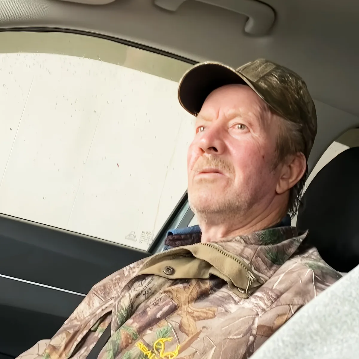 A man in a camouflage hat and jacket sits in a car, looking up.