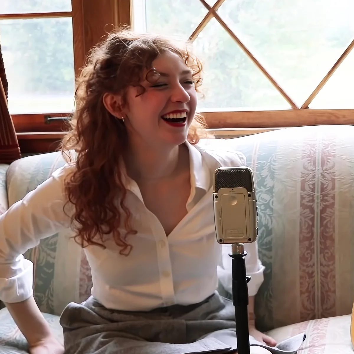 A woman with curly hair laughs while sitting on a patterned couch, with a microphone in front of her.