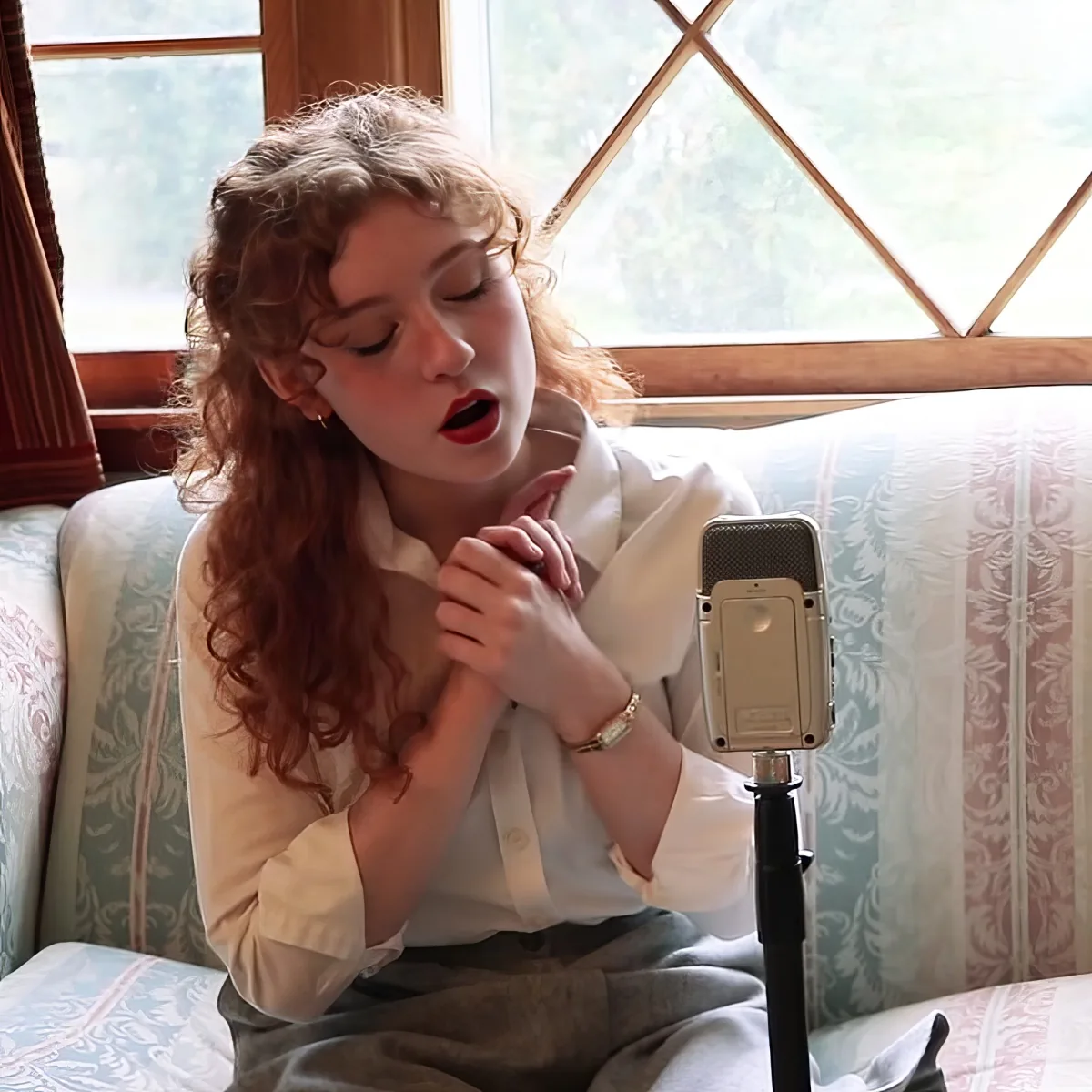 A woman with curly hair sings into a microphone, wearing a white shirt, with a patterned couch and window behind her.