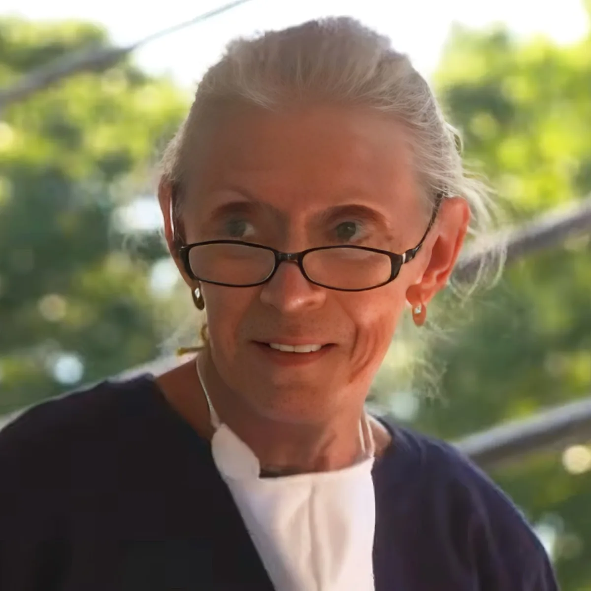 An older woman with white hair, glasses, and earrings, wearing a dark cardigan over a white shirt, smiles softly.