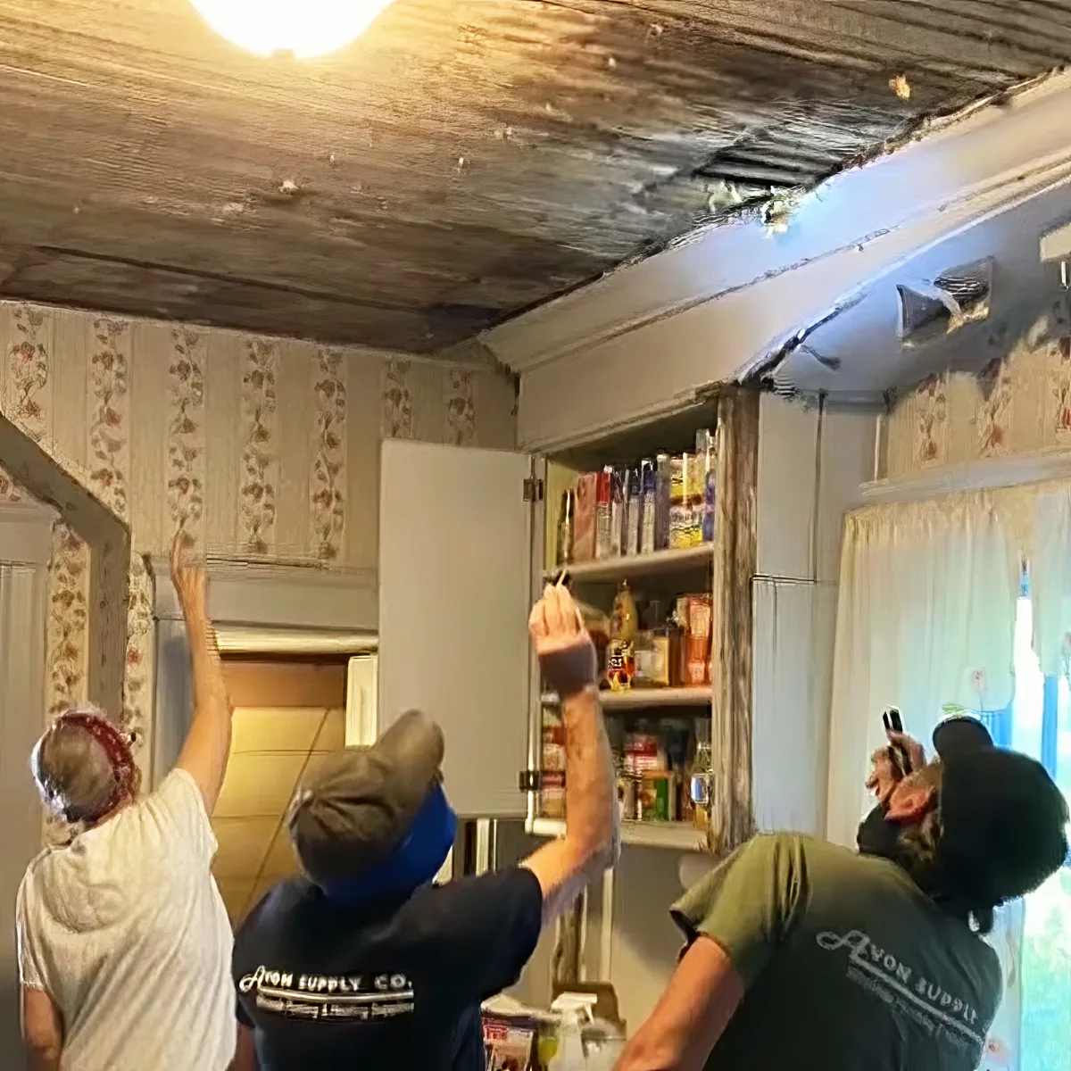 Three people in a kitchen with damaged ceiling and shelves, one pointing upwards.