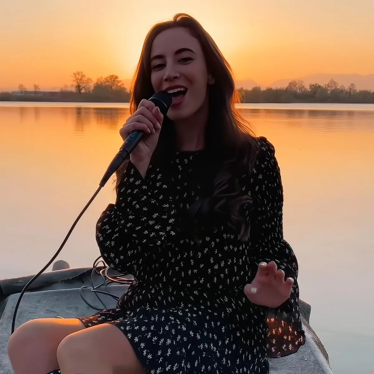 A woman in a floral dress sings into a microphone on a boat at sunset.